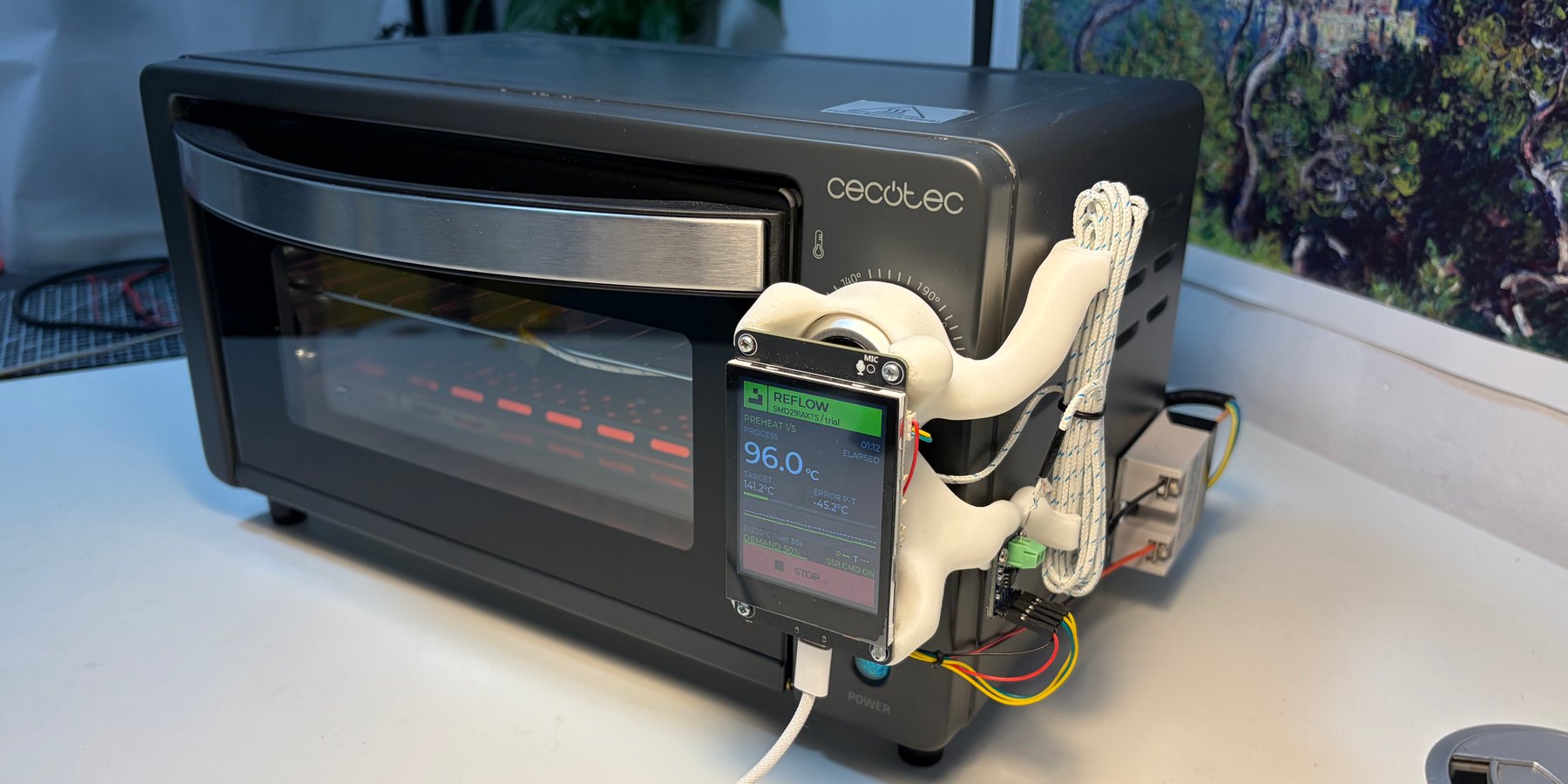The black oven mid-reflow, its quartz elements glowing through the glass door beside the touchscreen in its white printed bracket